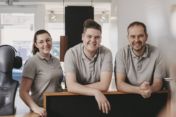 Drei freundliche Mitarbeiter in grauen Poloshirts stehen lächelnd am Empfangstresen eines Geschäfts.