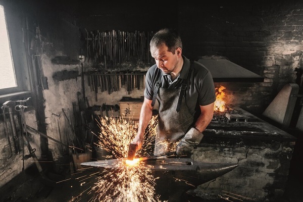 Ein Schmied bearbeitet glühendes Metall am Amboss, Funken fliegen in der Werkstatt.